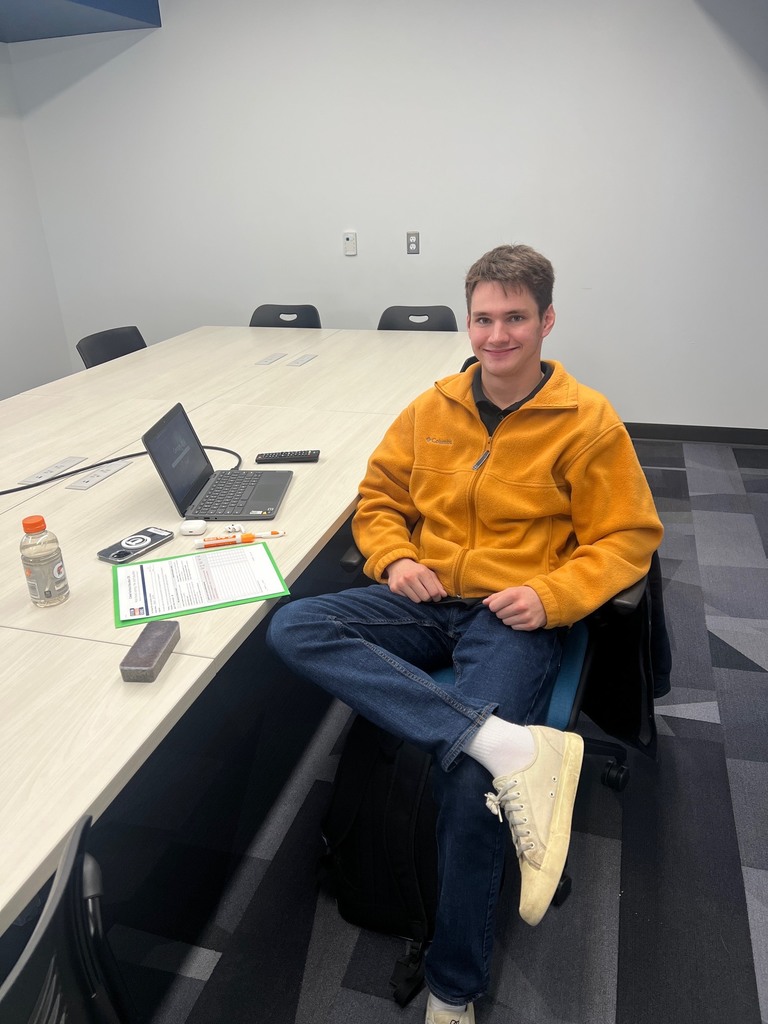 Student seated at a conference table in a meeting room, smiling at the camera. On the table are a laptop, clipboard with papers, pen, earbuds, phone, and a sports drink. The room has light-colored walls and modern office chairs.