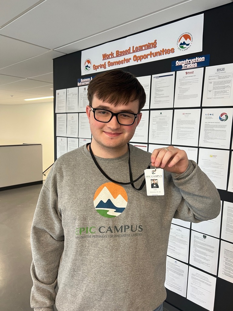 Student wearing a gray EPIC Campus sweatshirt holds up an EPIC Campus ID badge on a lanyard. The badge reads “EPIC Campus” and “Cole,” with “Intern” listed below. A bulletin board behind them displays “Work Based Learning Spring Semester Opportunities.”