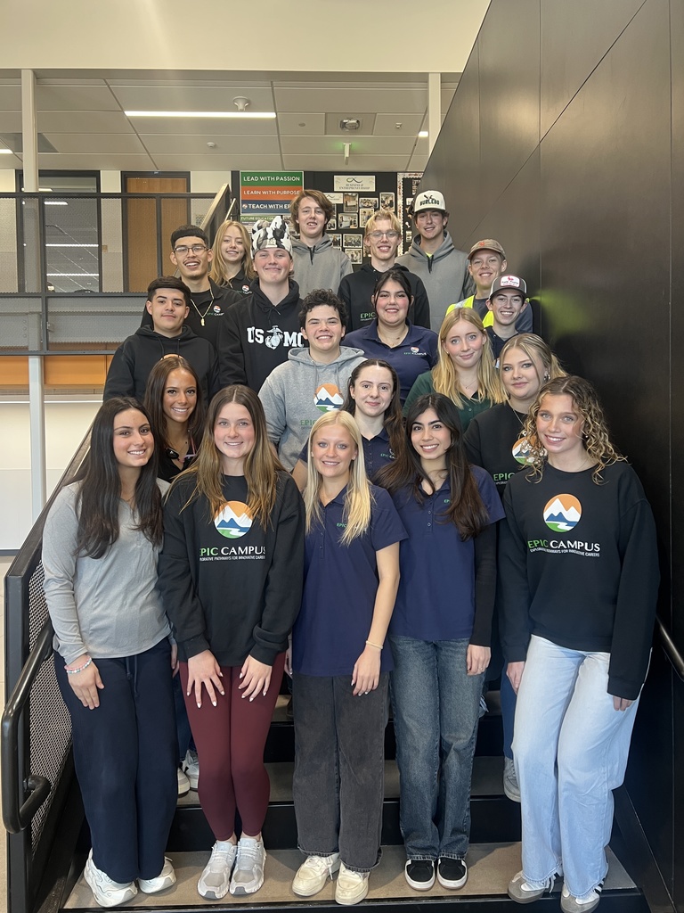 Large group of diverse high school students standing on indoor stairs at EPIC Campus, smiling at the camera. Many wear EPIC Campus shirts or hoodies with the mountain logo. A wall display behind them reads, “Lead with Passion, Learn with Purpose, Teach with EPIC.”