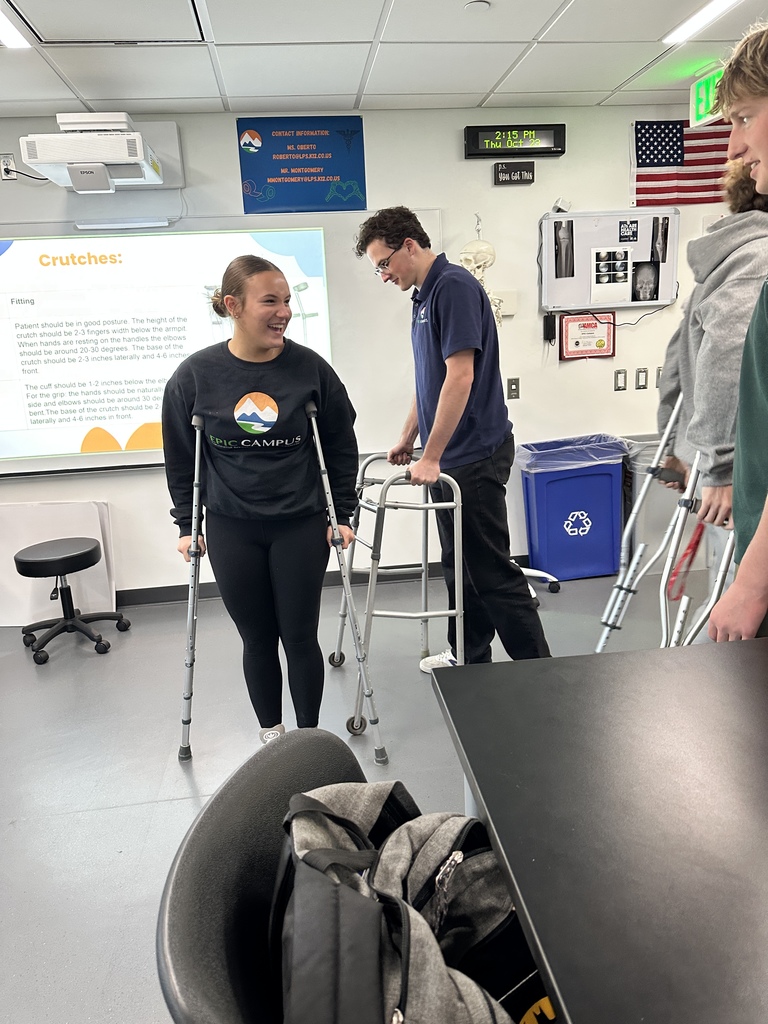 Students practice using mobility aids in a medical classroom. One student smiles while standing with crutches, and another uses a walker. Posters on the wall include instructions about using crutches.