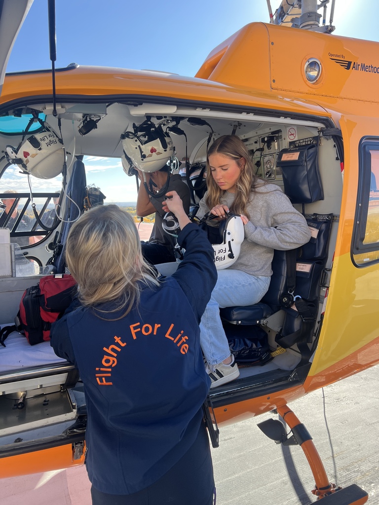 A student prepares to board a Flight For Life helicopter, assisted by a woman wearing a navy jacket with "Flight For Life" printed in orange on the back. Two students are seated inside the helicopter holding helmets.