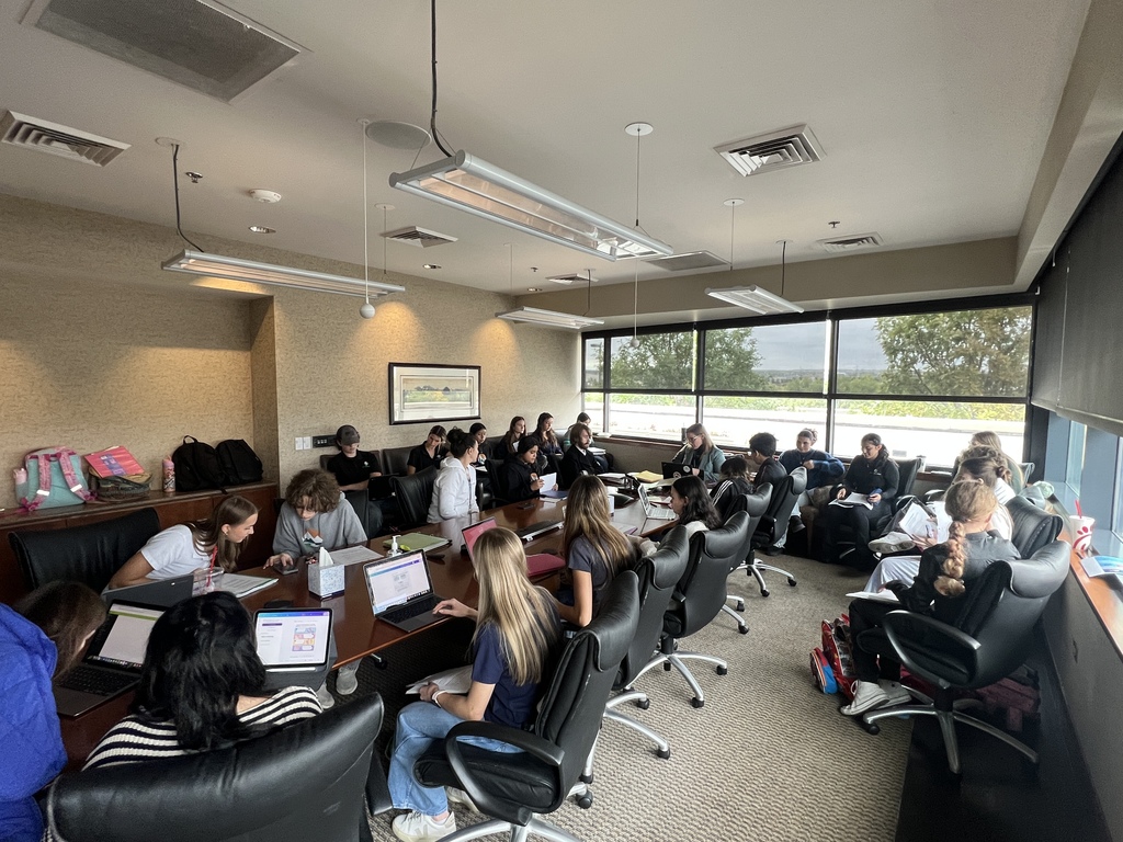 A group of students fill a large conference room, seated around a long table with laptops and notebooks. They appear engaged in collaborative work or a seminar, with backpacks and large windows visible in the background.