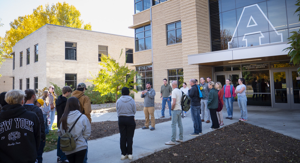 A group of high school students and adults stand outside a modern brick school building with large windows and a prominent white “A” displayed on the glass. A staff member addresses the group on a sidewalk surrounded by fall trees with bright yellow leaves. The entrance sign reads “Nutrein Agricultural Sciences Building.”