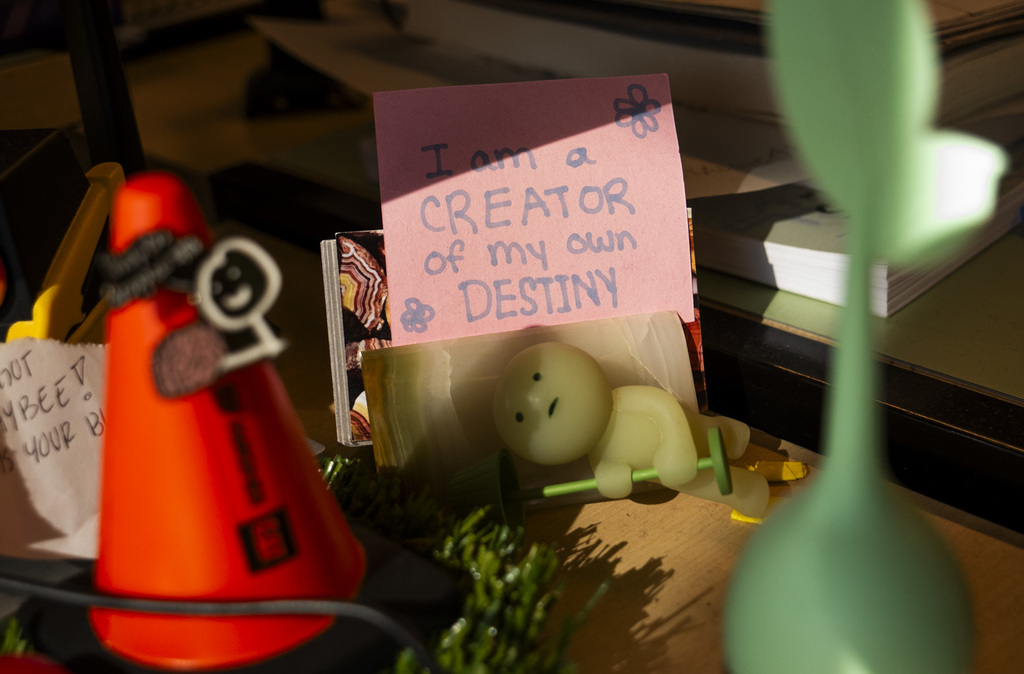 Close-up of a desk with a pink handwritten note that reads, “I am a CREATOR of my own DESTINY.” Nearby are small decorative items, including a green figurine holding a barbell, a red toy cone, and stacks of paper in warm sunlight.