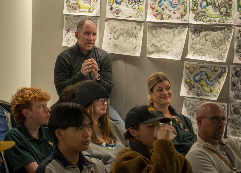 Students and adults sit closely together indoors, listening attentively. Landscape design sketches are pinned across the wall behind them, showing detailed garden and park layouts. The group represents a mix of genders and ethnic backgrounds, dressed in casual school attire.