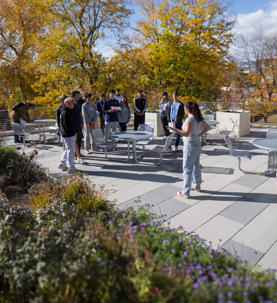 A group of high school students stand on an outdoor rooftop patio with metal tables and chairs, listening to a student speaker. Fall trees with bright yellow leaves fill the background, and landscaped planters with flowers and greenery line the foreground.