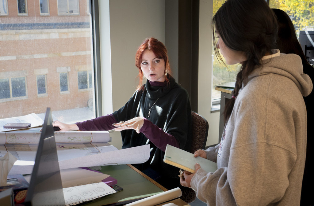 Two high school students sit by a window reviewing large landscape design drawings. One student gestures while explaining details on the plan, and the other holds a notebook. Sunlight streams in, illuminating the workspace and highlighting the detailed design papers spread across the desk.