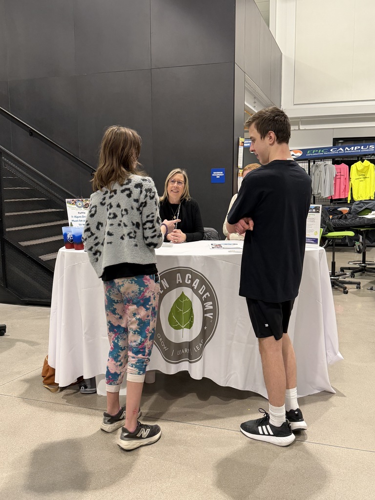 Two students speak with a representative at a booth labeled “Green Academy,” featuring a circular leaf logo and the words “Grow | Learn | Lead.” The representative sits behind a white tablecloth with informational handouts displayed.