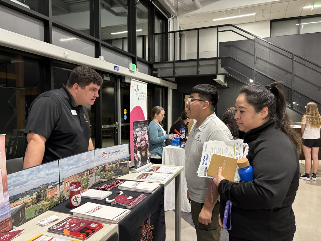 A representative from Colorado Mesa University speaks with a student and an adult at a college fair table inside a modern school commons area. The table displays brochures, pens, a “Colorado Mesa University” sign, and a red Maverick mascot graphic. Other booths and attendees are visible in the background near large windows and a staircase.