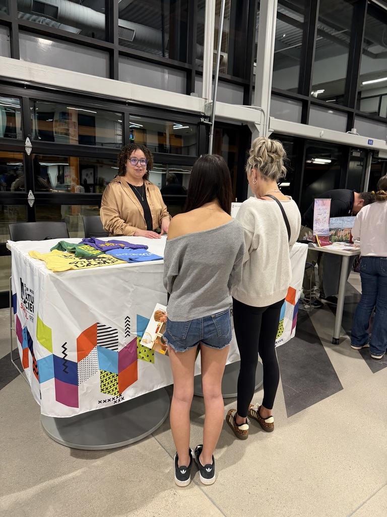 Two students speak with a representative behind a table covered with colorful T-shirts and informational materials. The tablecloth features a bright geometric pattern, and additional college booths are set up nearby in the large indoor commons.