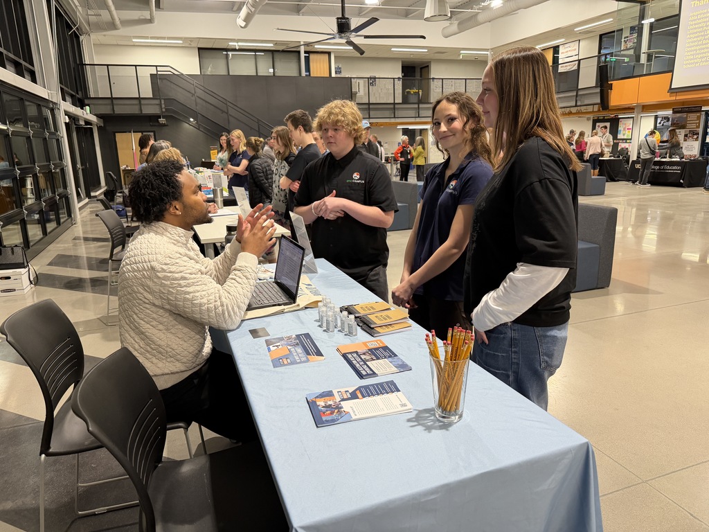 Three EPIC Campus high school students stand at a booth speaking with a representative seated behind a table covered with brochures and pencils. The open commons space includes multiple college tables and attendees mingling in the background.
