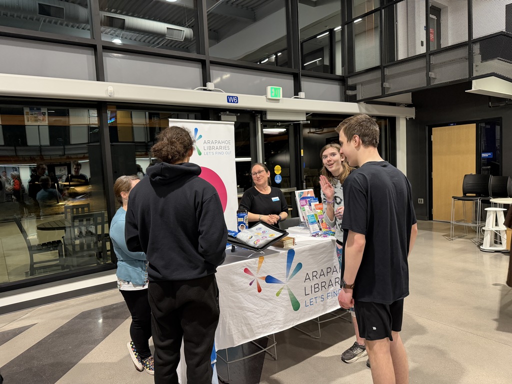 Students gather at an Arapahoe Libraries booth with a white tablecloth displaying the Arapahoe Libraries logo and the phrase “Let’s Find Out.” A staff member shares information and materials while students listen and look at brochures and a display binder.