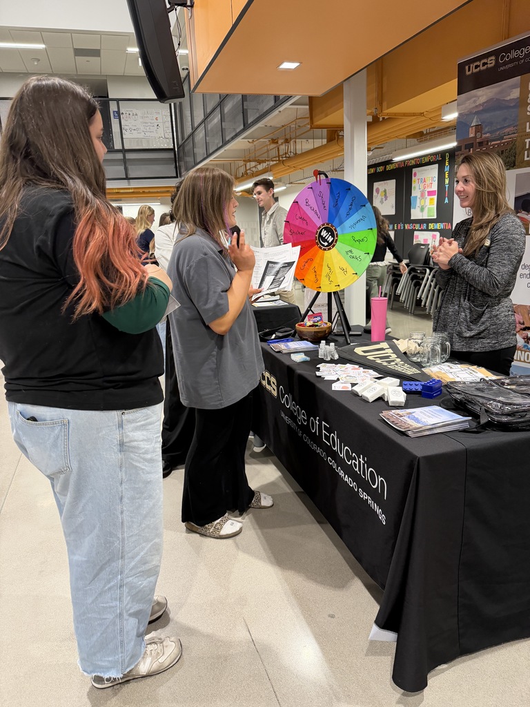 An MSU Denver School of Education representative speaks with a small group at a booth featuring a blue tablecloth and a vertical banner reading “MSU Denver School of Education.” A colorful prize wheel and informational materials are displayed on the table.
