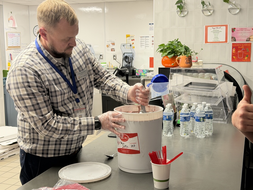 An EPIC Campus staff member wearing gloves scoops ice cream from a large white tub labeled “Junie” behind a stainless-steel counter in a bright school café space. Red spoons, small sample cups, bottled water, and countertop equipment are arranged nearby, with colorful menu signs posted on the tiled wall.