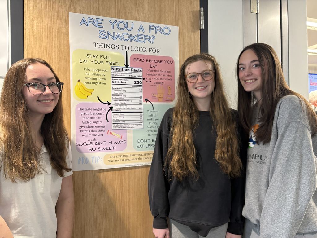 Three high school students stand smiling in front of a classroom door next to a colorful poster titled “Are You a Pro Snacker? Things to Look For.” The poster includes nutrition tips such as “Stay Full – Eat Your Fiber!” and “Read Before You Eat,” along with a sample Nutrition Facts label and reminders that “Sugar Isn’t Always So Sweet.”