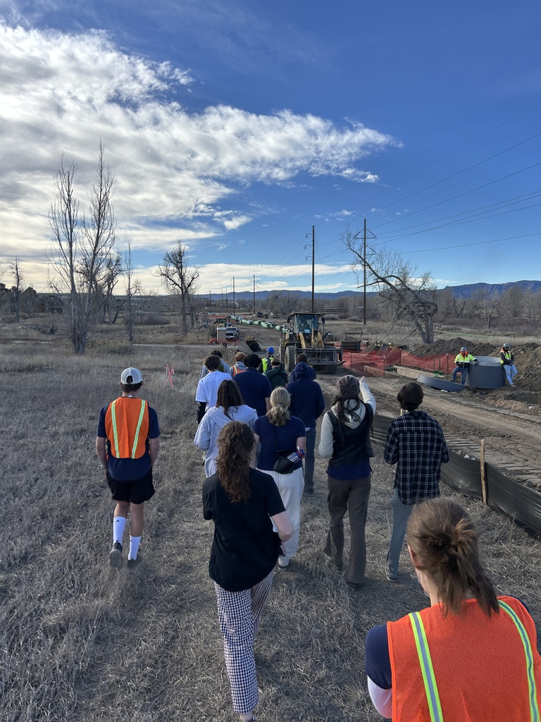 Students walk through a grassy open field toward a construction area with heavy equipment and large green pipes. Several individuals wear bright orange safety vests, and mountains and power lines are visible under a blue sky.