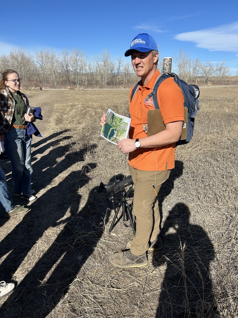 An outdoor guide wearing an orange polo shirt and a blue “South Suburban” hat holds a map and speaks to students in a grassy field. A backpack and spotting scope stand nearby, with leafless trees and open space in the background.