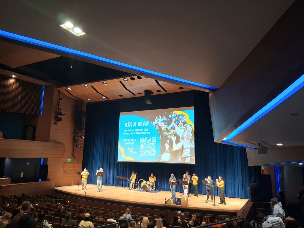 A school band performs on stage in a large auditorium with blue curtains and wood paneling. A projected slide above them reads “ASK A BEAR by Future Teacher Club @unco_futureteacherclub” with a QR code and a photo of cheering students. Audience members sit in theater-style seats watching the performance.