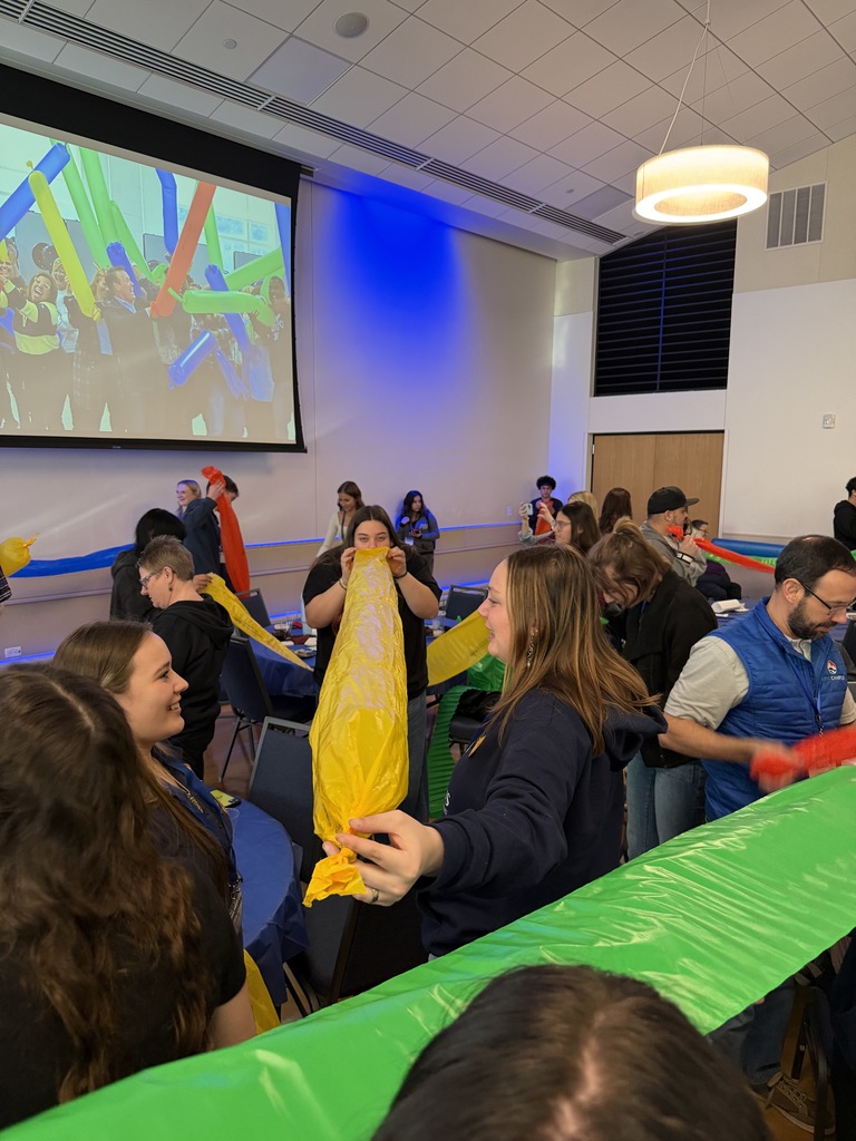 Students and staff participate in a hands-on activity in a conference room, holding long colorful inflatable tubes. A large screen in the background displays an image of people celebrating with similar balloons, adding to the energetic atmosphere.