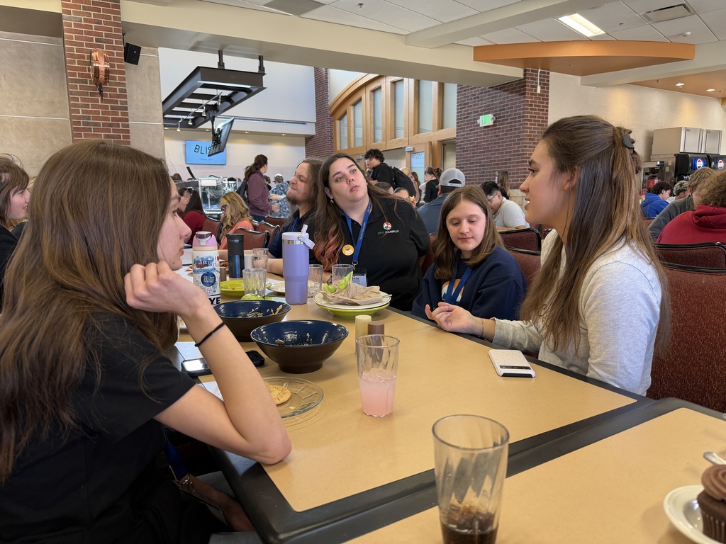 A small group of students and an EPIC Campus staff member sit together at a dining hall table, talking and eating. Plates, bowls, drinks, and water bottles are spread across the table in a busy campus café setting.