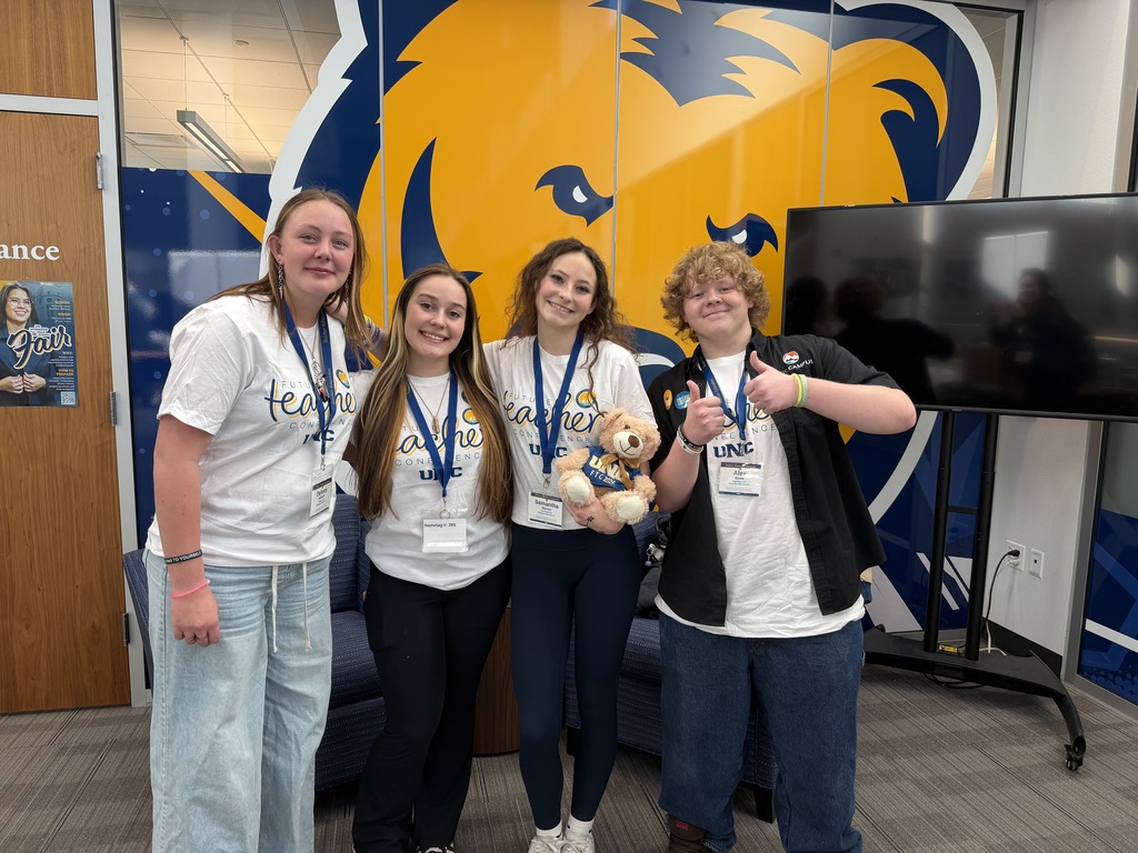 Four students stand smiling in front of a large blue and gold UNC bear graphic. They wear “Future Teacher Conference UNC” T-shirts and name badges, and one student holds a small teddy bear mascot while another gives a thumbs-up.