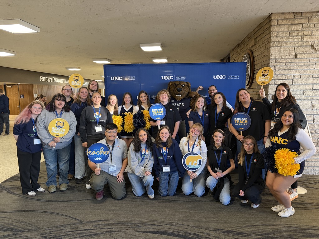 A large group of students and staff pose together in front of a blue “UNC – University of Northern Colorado” backdrop with the UNC bear mascot. Some hold signs reading “Dream Teach Inspire” and “Bear to Inspire,” and a cheerleader with gold pom-poms stands at the side of the group.