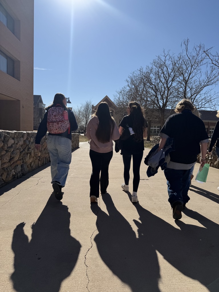 Four high school students walk across a sunny college campus pathway between brick buildings and stone walls. Long shadows stretch across the sidewalk under a clear blue sky.