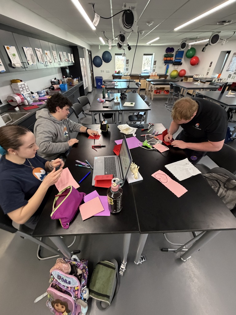 Three high school students work together at black lab tables in a classroom, making Valentine’s cards with colorful paper and markers. A laptop, water bottle, scissors, and craft supplies are spread across the table. The students, dressed in casual school clothing, concentrate on writing and decorating. The classroom includes cabinets with anatomical models, exercise balls mounted on the wall, and overhead equipment, suggesting a health or science lab setting.