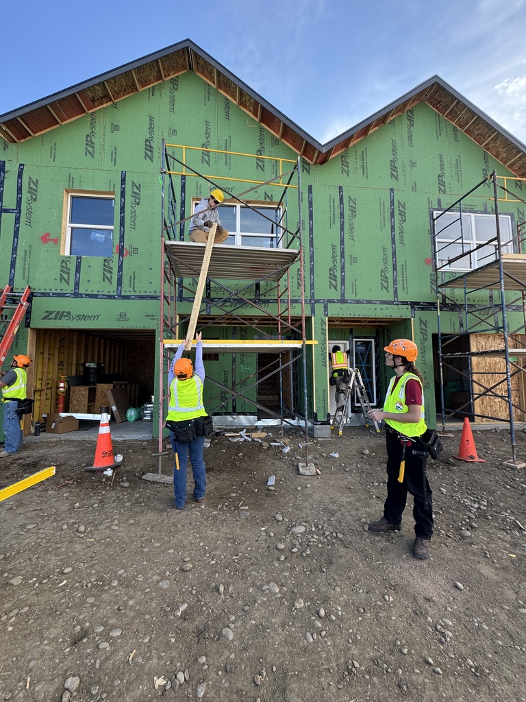 Students in hard hats and high-visibility vests work on scaffolding in front of a two-story house under construction covered in green ZIP System sheathing. One student kneels on the scaffold platform while another hands up a long board from the ground. Additional students stand nearby observing and assisting. Ladders, traffic cones, and construction materials surround the site under a clear blue sky.