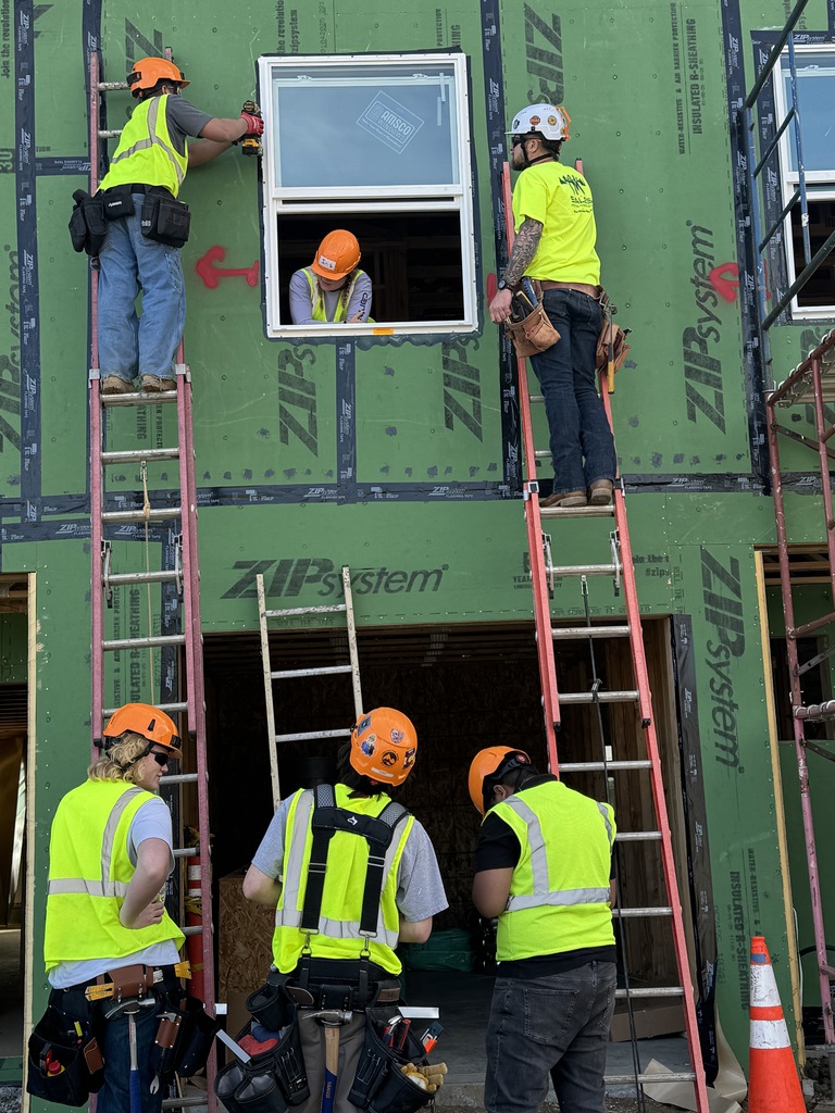 A group of construction students wearing orange hard hats and neon safety vests install a window on the exterior of a house framed with green ZIP System panels. Two students stand on ladders securing the window with tools while another leans out from inside the opening. Three classmates stand below watching and discussing the process. A traffic cone and scaffolding are positioned near the entryway.