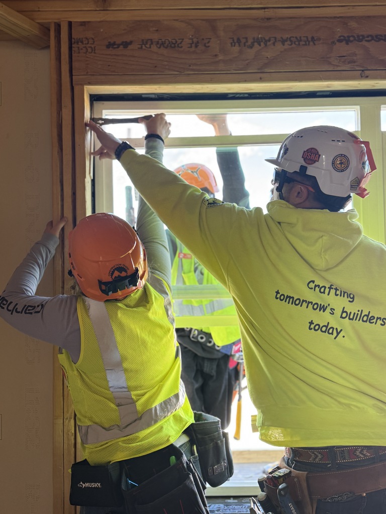 Two construction students wearing bright safety vests and hard hats install a window frame inside a partially finished building. One student holds a pry bar at the top of the frame while another steadies the side. The student on the right wears a neon hoodie with the text “Crafting tomorrow’s builders today.” printed on the back. A third person in safety gear is visible outside through the window opening.