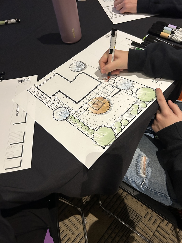 A student uses a marker to add color to a residential landscape design plan featuring a house layout, patio, pathways, trees, and shrubs. Green and orange shading highlights different landscape elements. Additional printed plan sheets labeled “RESIDENCE” and “Designscapes Colorado” are visible on the table, along with a pink water bottle and a set of markers arranged nearby.