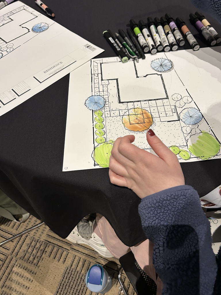A student colors a detailed landscape design plan on white paper at a black classroom table. The drawing shows a house footprint, walkways, trees shaded in blue and green, shrubs, and a patio area colored orange. A row of assorted markers is lined up at the top of the table. The printed page includes the text “RESIDENCE” and a “Designscapes Colorado” logo in the corner.
