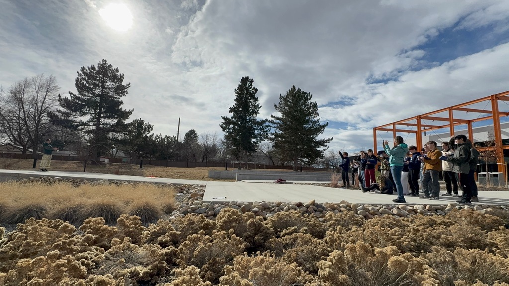 A group of high school students stand outdoors on a concrete patio observing a small launch demonstration in a landscaped courtyard. An instructor stands at a distance on the left, while students on the right watch and hold clipboards. Dry grasses, rocks, evergreen trees, and an orange metal pergola frame the scene under a bright, partly cloudy sky.