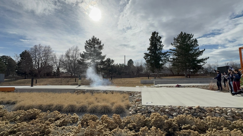 Students watch as a small rocket launches, producing a cloud of white smoke on a concrete walkway in an outdoor courtyard. An instructor stands nearby supervising while students observe from a safe distance with clipboards. The area includes tiered concrete seating, river rocks, native grasses, and tall trees beneath a sunny sky.
