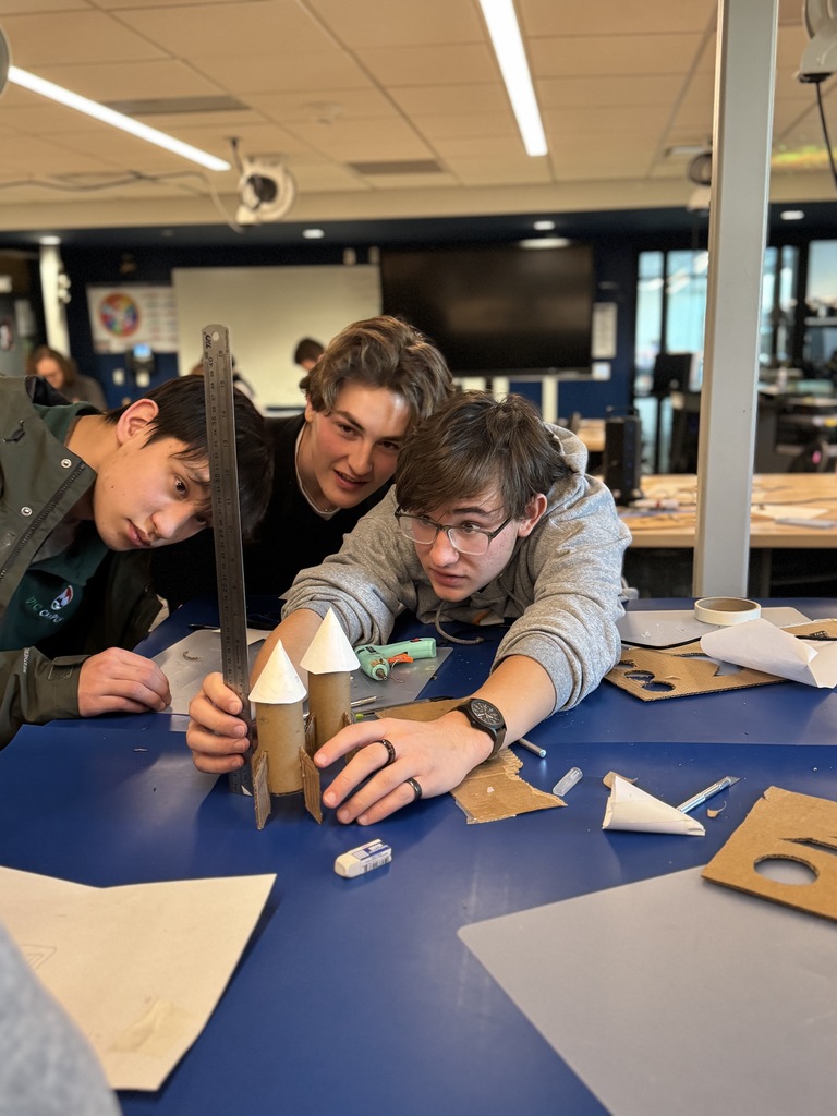 Three high school students lean over a table in a STEM classroom, carefully measuring a small cardboard rocket model with a metal ruler. The model is made from cardboard tubes and paper cones. Craft supplies, scissors, and tools are scattered on the blue tabletop as the students focus on precise measurements.