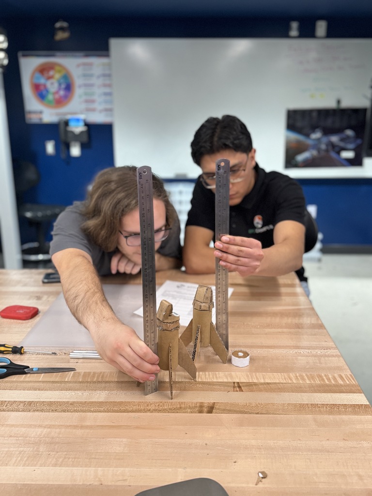 Two high school students measure cardboard rocket models on a wooden workbench in a classroom lab. Each student holds a metal ruler vertically next to the model to check height and alignment. Scissors, tape, and tools are arranged on the table, with a whiteboard and classroom displays visible in the background.