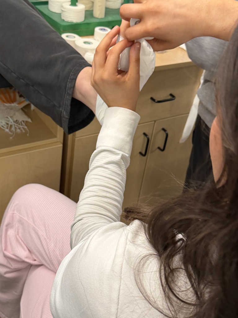 A close-up view shows a student carefully wrapping white athletic tape around a classmate’s ankle and heel. The student’s hands secure the tape snugly while the classmate’s leg rests on a padded table. Multiple rolls of tape are visible in the background.