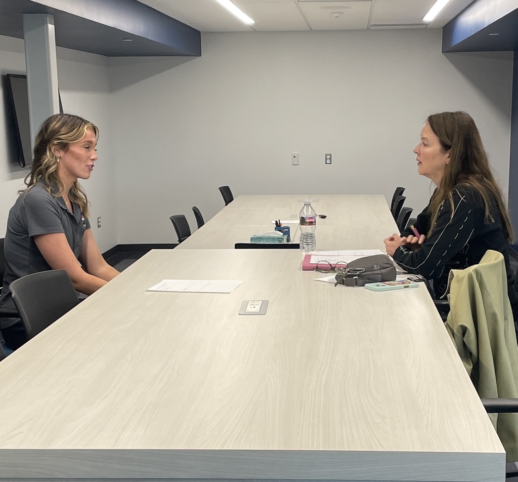 Two adult women sit across from each other at a long conference table engaged in conversation, suggesting a mock interview or mentoring session. Papers and a water bottle rest on the table between them in a small meeting room with neutral walls and overhead lighting.