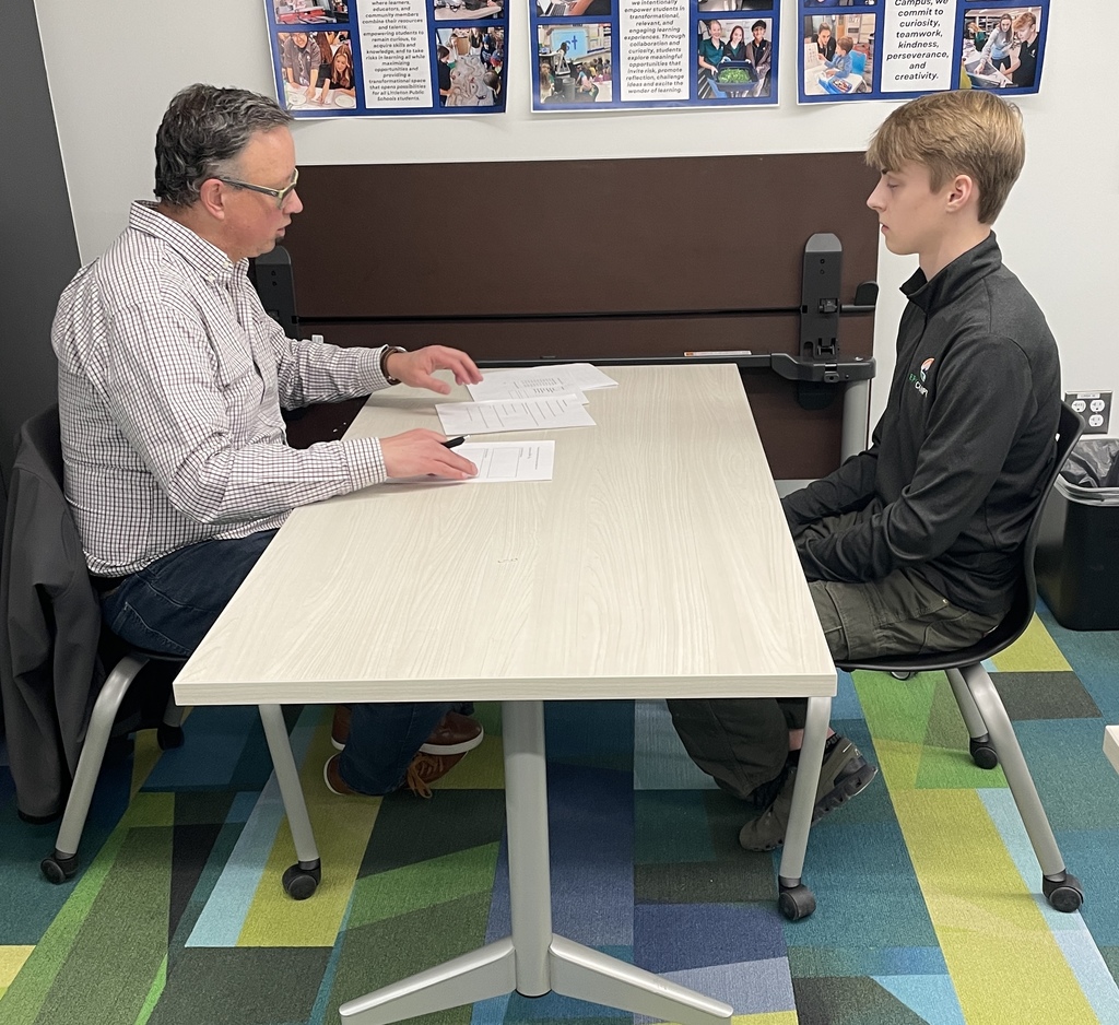 An adult man reviews paperwork with a high school student seated across from him at a small table in a classroom. The student listens attentively while the adult gestures toward printed documents, simulating an interview or professional meeting. Posters highlighting school programs are displayed on the wall behind them.