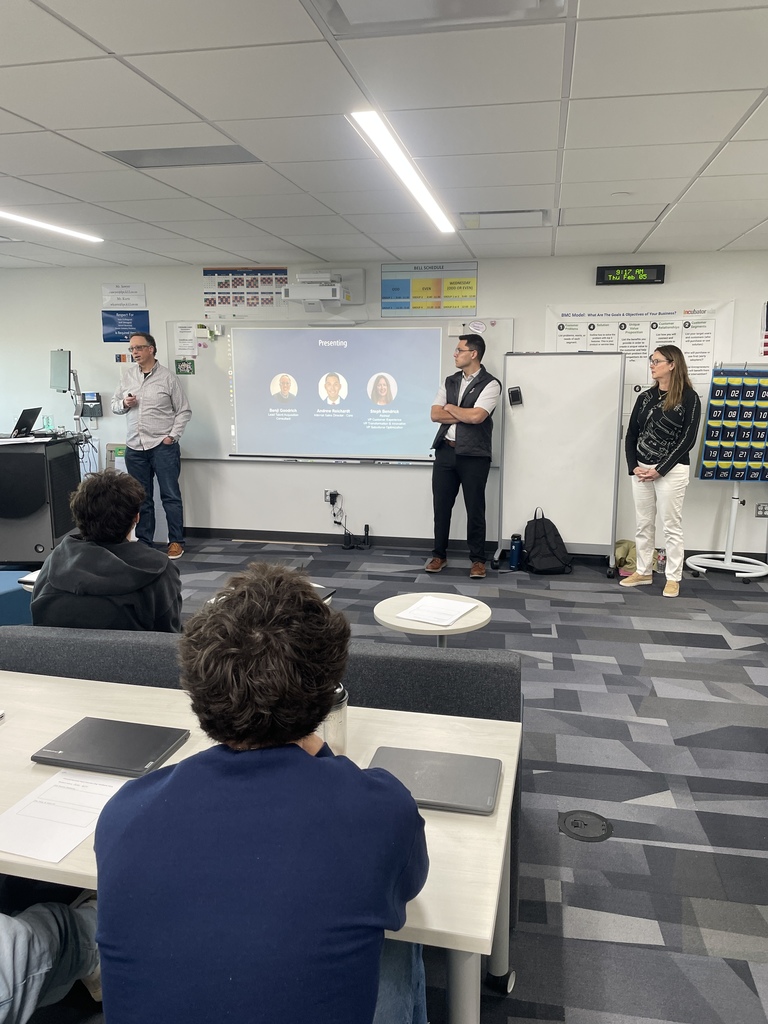 Three adult presenters stand at the front of a classroom speaking to seated high school students. A projected slide behind them reads “Presenting” and displays three headshots with names and titles. Students sit at tables with laptops while listening. Classroom posters, a whiteboard, and a digital clock are visible on the walls.