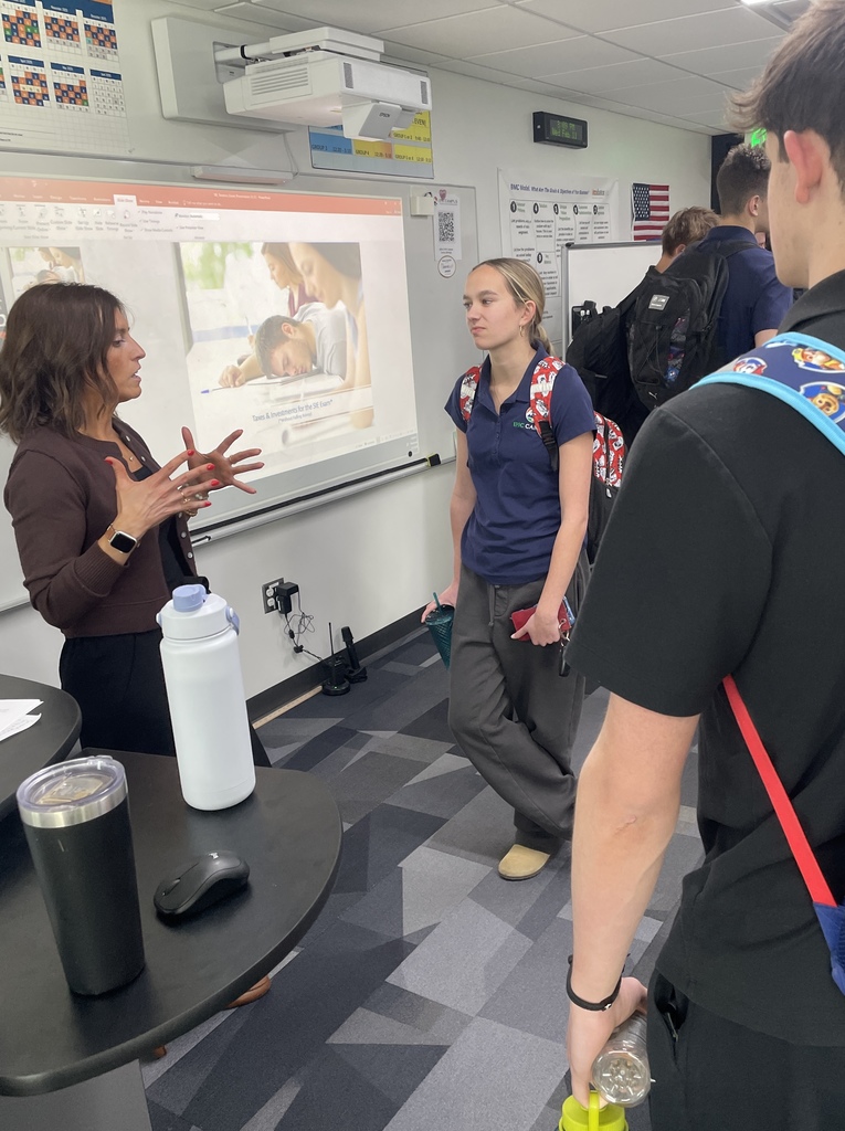 A guest speaker talks with a small group of high school students at the front of the classroom after a presentation. The slide behind them reads “Taxes & Investments for the 21st Century” and shows a student studying. The students, wearing backpacks and school apparel, listen attentively while standing near the projection screen and classroom whiteboard.