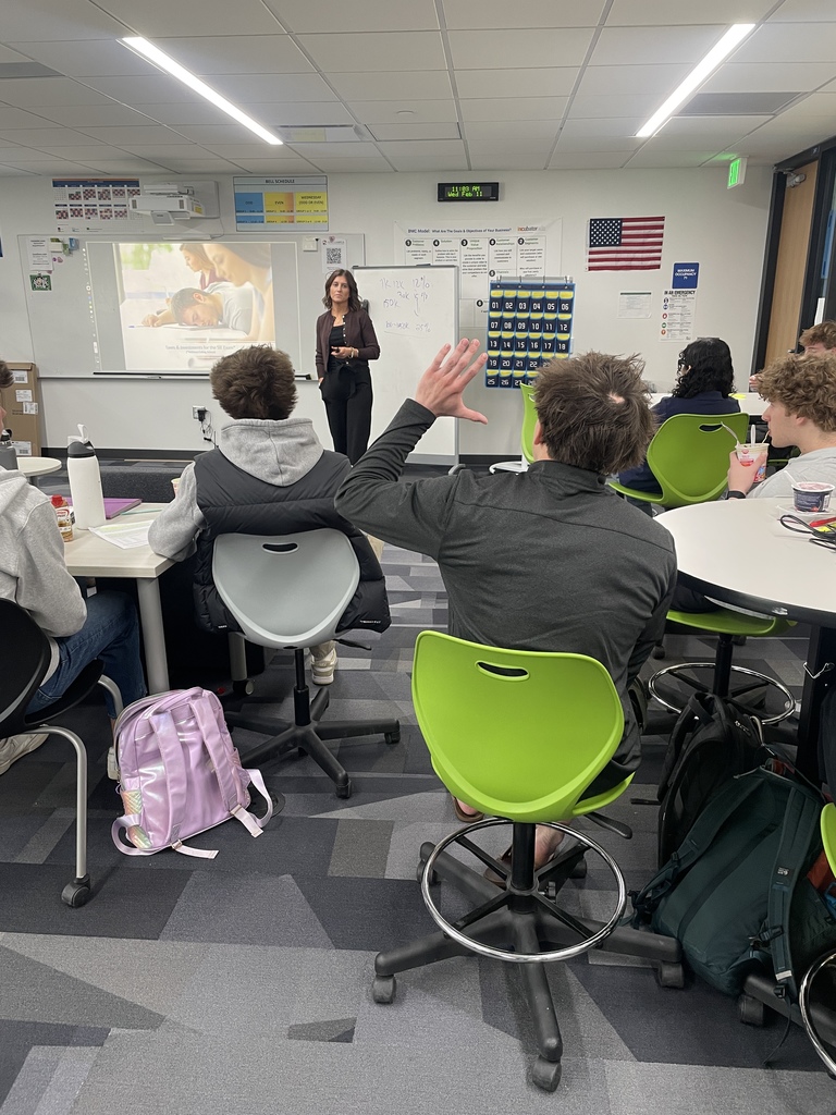 A female guest speaker stands at the front of a classroom presenting to seated high school students. A projected slide behind her shows an image of a student studying with the title “Taxes & Investments for the 21st Century.” One student raises his hand while others sit at round tables with notebooks and water bottles. An American flag, whiteboard notes, and a digital clock are visible on the wall.
