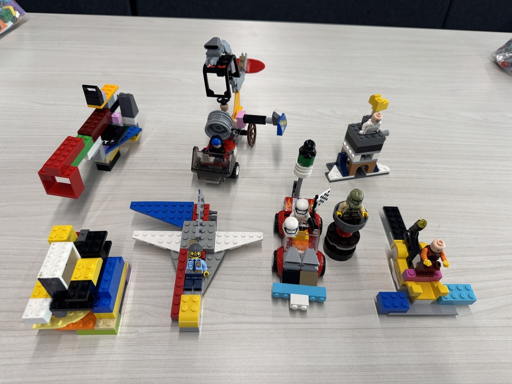 Several small LEGO creations are arranged on a classroom table, including colorful vehicles, a small airplane with a pilot figure, a cart with accessories, and multiple minifigures. The builds feature bright red, blue, yellow, and white bricks and include characters such as a police officer and helmeted figures, suggesting creative design or engineering activities.