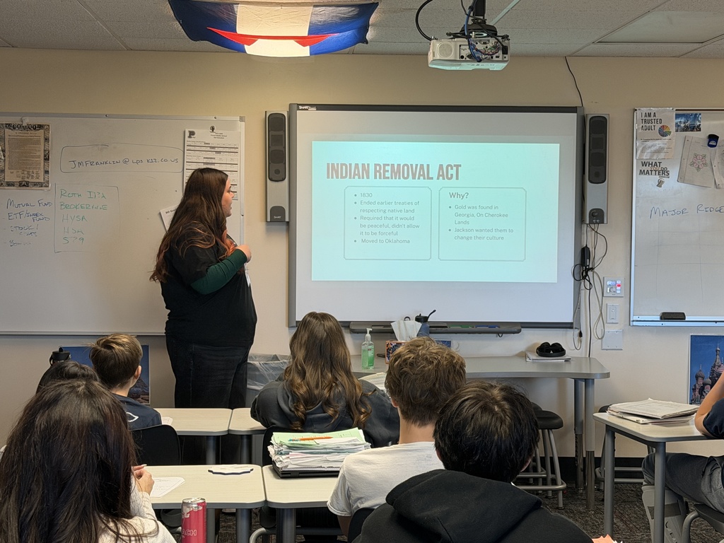 A teacher presents a slide titled “Indian Removal Act” to a class of high school students seated at desks. The slide includes bullet points about 1830, Oklahoma, and reasons related to gold in Georgia and Cherokee lands.