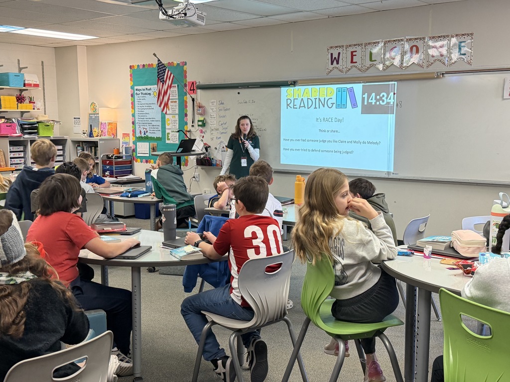 An elementary classroom during shared reading time shows a teacher standing near a screen that reads “Shared Reading – It’s RACE Day!” Students sit at round tables with books and water bottles, listening and preparing to discuss.