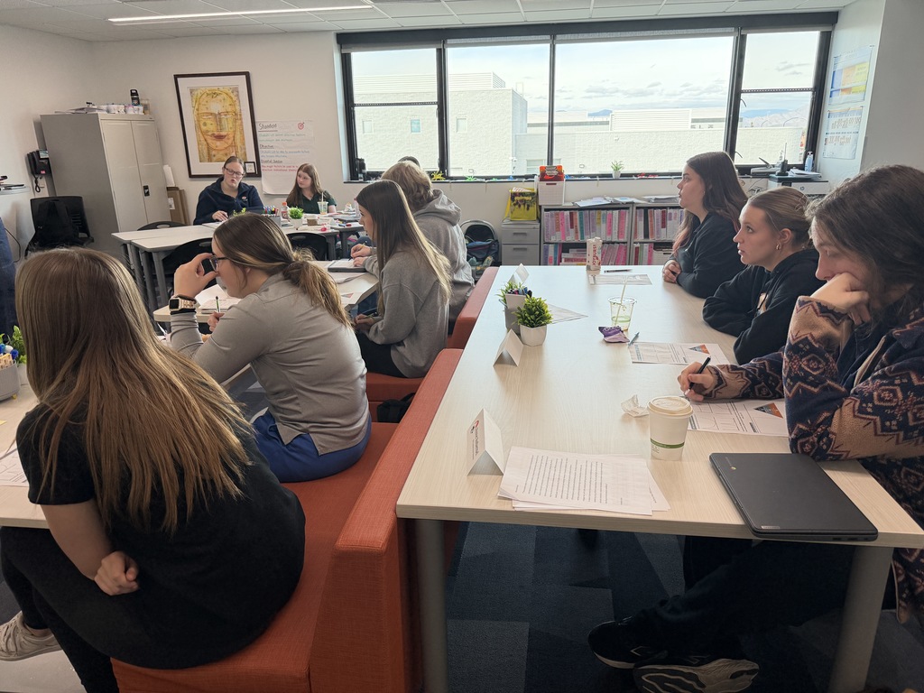 An instructor stands at the front of a classroom pointing to projected graphs on a whiteboard while adult participants sit at tables facing forward. An American flag and a “Strong Teachers, Epic Classrooms, Future Educators” banner hang on the wall.