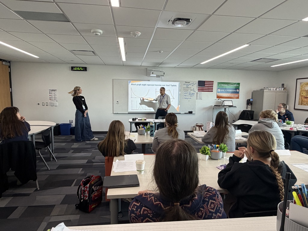 Adult learners sit at tables in a bright classroom with large windows, listening and taking notes during a professional development session. Papers, name tents, coffee cups, and laptops are on the tables, and binders line a shelf beneath the windows.