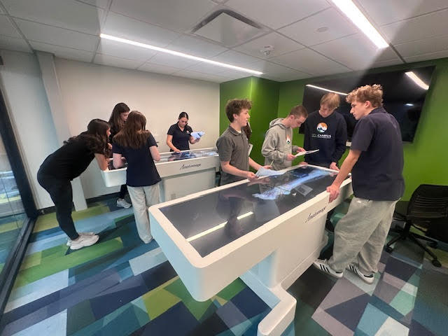 A wider view of a classroom shows two groups of high school students gathered around separate white Anatomage tables. The tables display full-length digital human anatomy images. Students of diverse backgrounds review printed worksheets and collaborate while interacting with the touchscreens. The room includes green and white walls, modern furniture, and geometric-patterned carpet flooring.