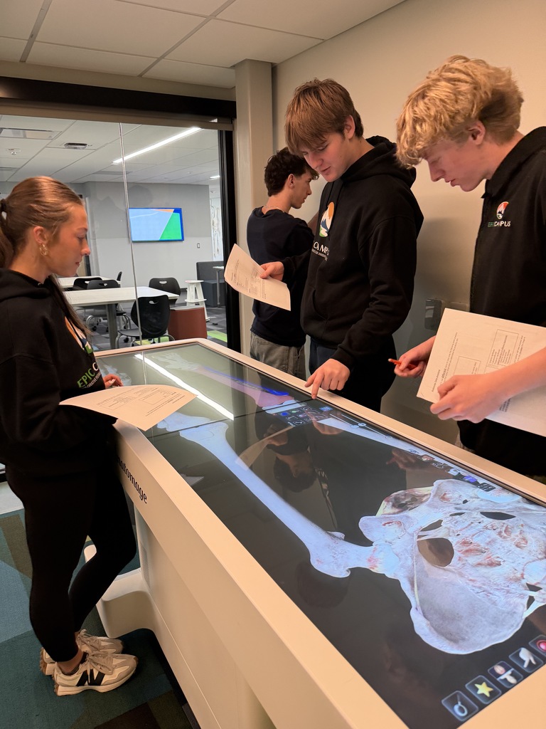 Four high school students wearing school-branded hoodies and casual attire stand around an Anatomage table displaying a 3D image of a human pelvis and upper leg bones. One student points to on-screen controls while others review worksheets and discuss the image. The setting is a modern classroom with glass walls, flexible seating visible in an adjacent room, and patterned carpet flooring.